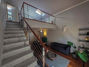Eine Treppe mit hölzernem Geländer führt zu einer Zwischenebene, einem dunkelgrauen Sofa, einem runden Couchtisch und einem Blumentopf.