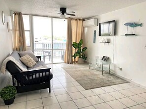 Ein Wohnzimmer mit einer schwarzen Couch, einem Topf mit Pflanze, einem Glastisch und einem an der Wand befestigten Fernseher.