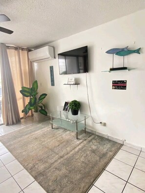 Ein modernes Wohnzimmer mit Glastisch, flachbildfernseher an der Wand, Blumentopf und Schild mit Hinweis auf kostenloses WLAN.