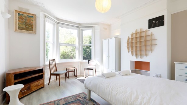 Ein Schlafzimmer mit einem großen Bett, einem Schreibtisch, einem Stuhl, einem Kleiderschrank und einem Fenster mit Blick auf Bäume.