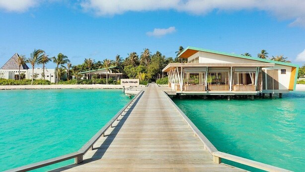 Resort com píer de madeira que leva a bangalôs sobre a água, águas turquesa cristalinas e palmeiras.