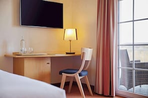 Habitación de hotel con cama, escritorio con lámpara, silla, televisión y ventana con cortinas.