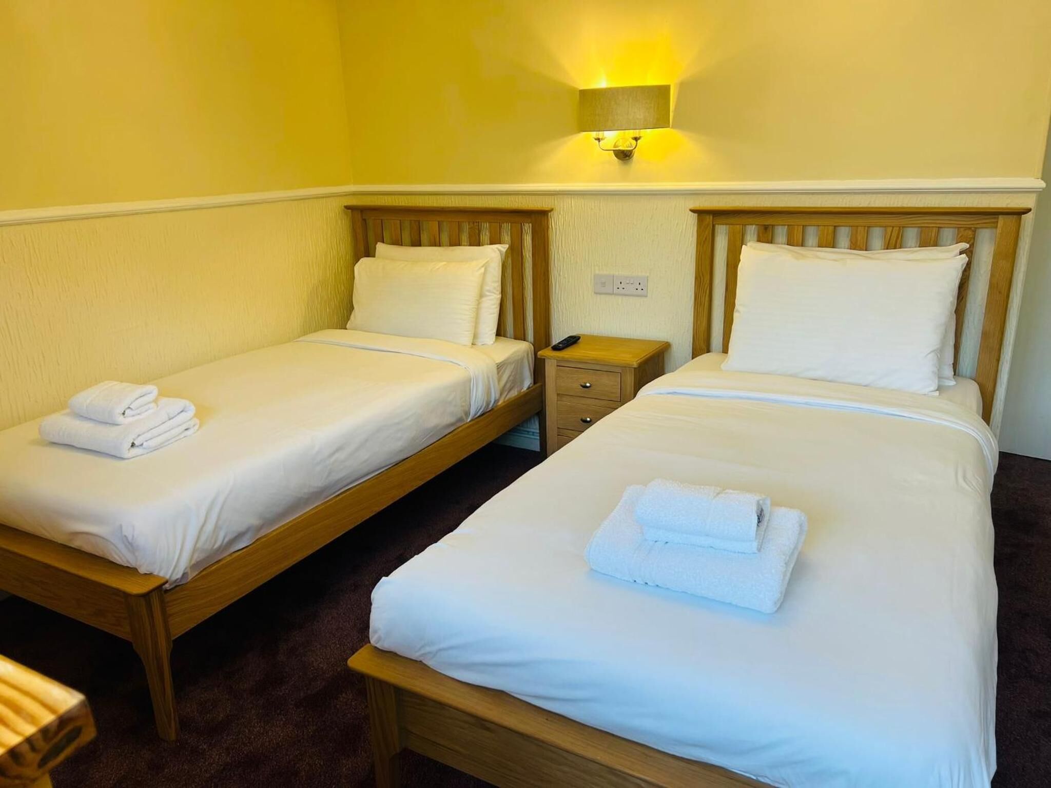 A hotel room with two single beds, each with a wooden headboard, white bedding, and a folded towel on the bed. There is a nightstand with a lamp between the beds.
