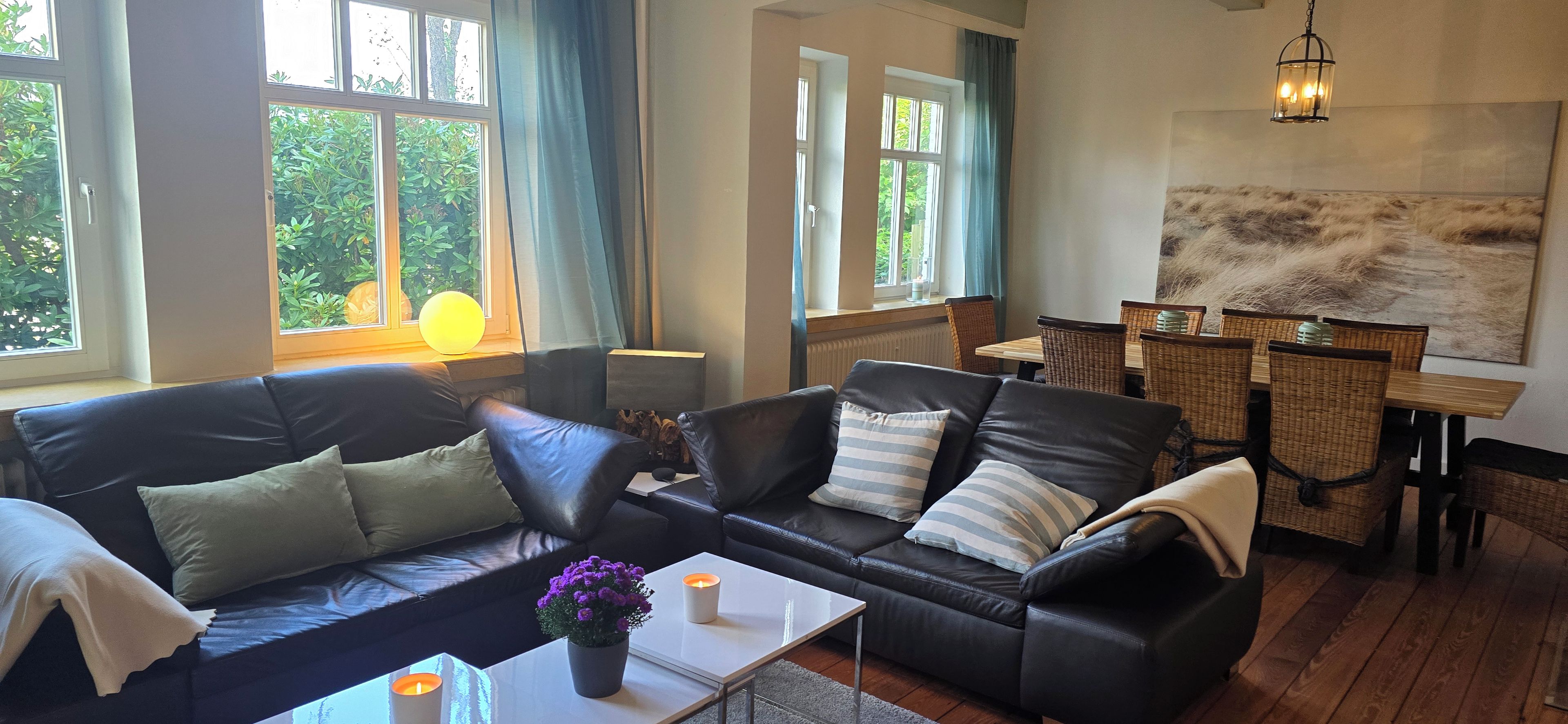 A living room with a black leather sofa, a dining table with wicker chairs, and a large window with curtains.