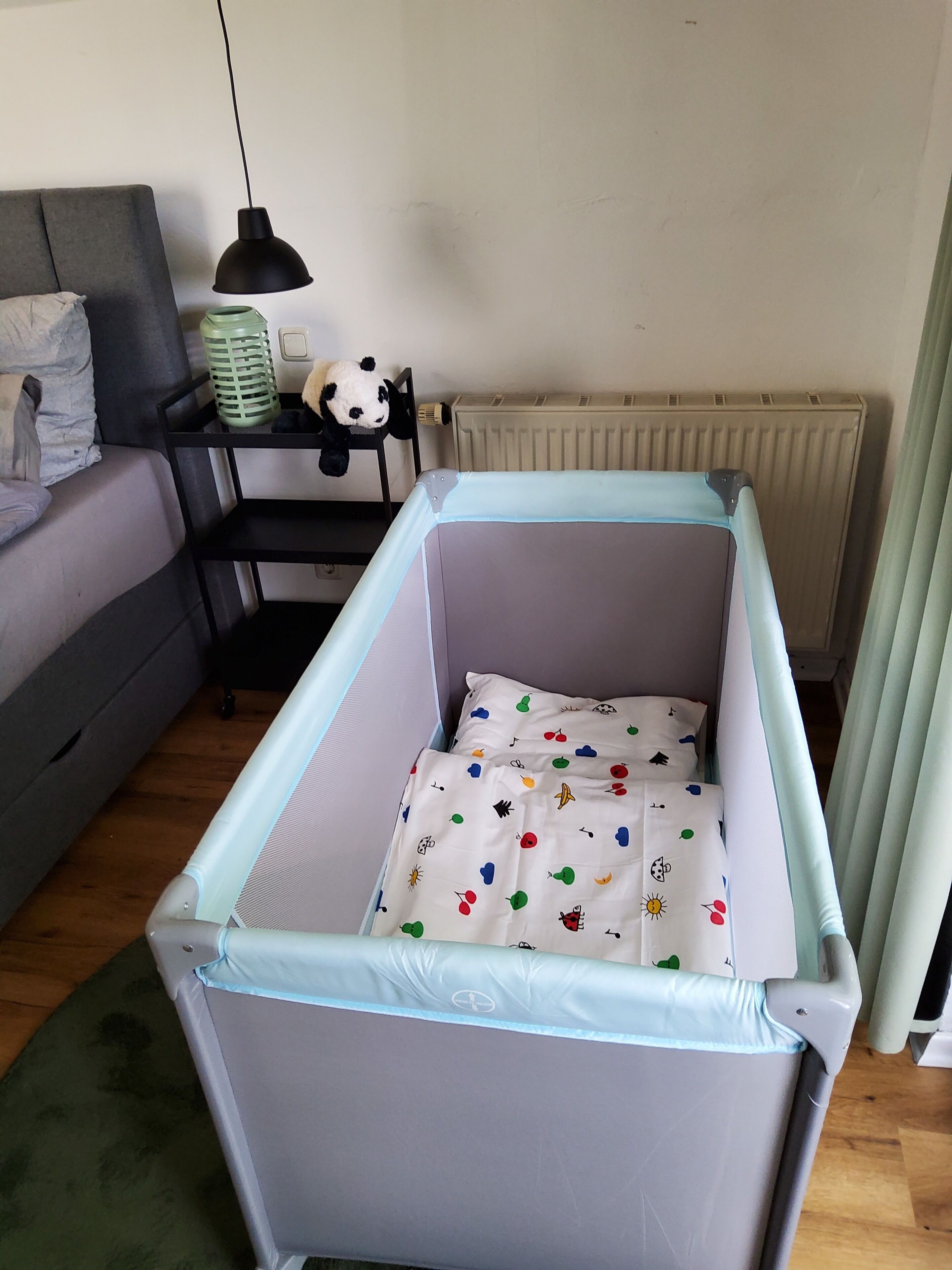 Ein Babybett mit gemusterten Matratze, ein graues Sofa, ein schwarzer Nachttisch mit Lampe und eine Heizung.
