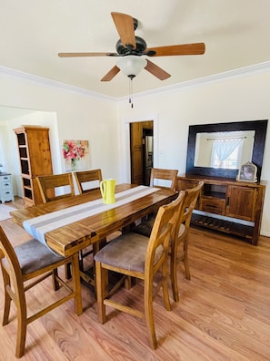 A dining area with a wooden table, chairs, a ceiling fan, and a kitchen visible through an open doorway.