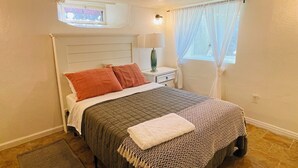 Un dormitori amb un llit, una tauleta de nit, una làmpada, una finestra amb cortines, un petit fanal i una catifes.