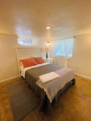 A bedroom with a bed, a nightstand, a lamp, a window with curtains, a small skylight, and a rug.
