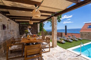 Eine Steinterrasse mit Holztisch und -stühlen, einem Pool und Blick auf das Meer.