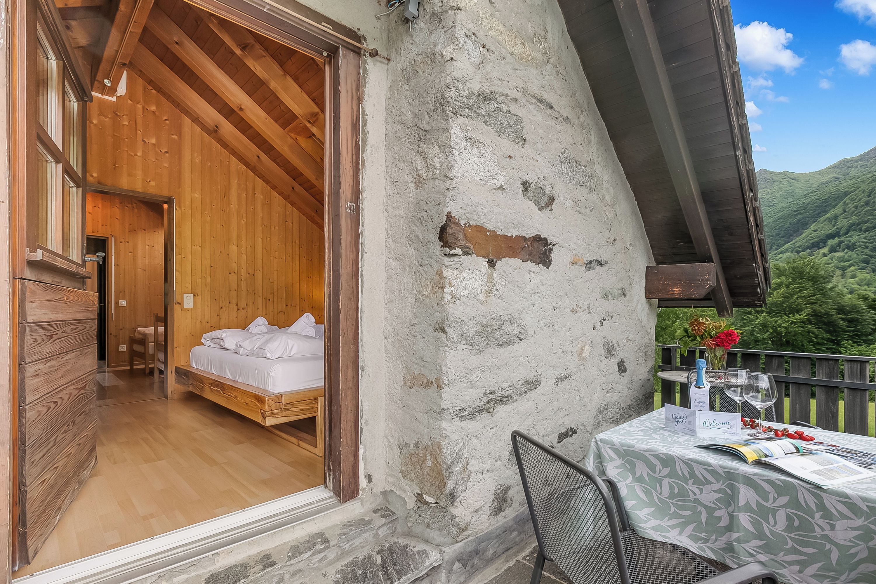 Una habitación de madera con una cama, un balcón con mesa y vistas a un paisaje montañoso.