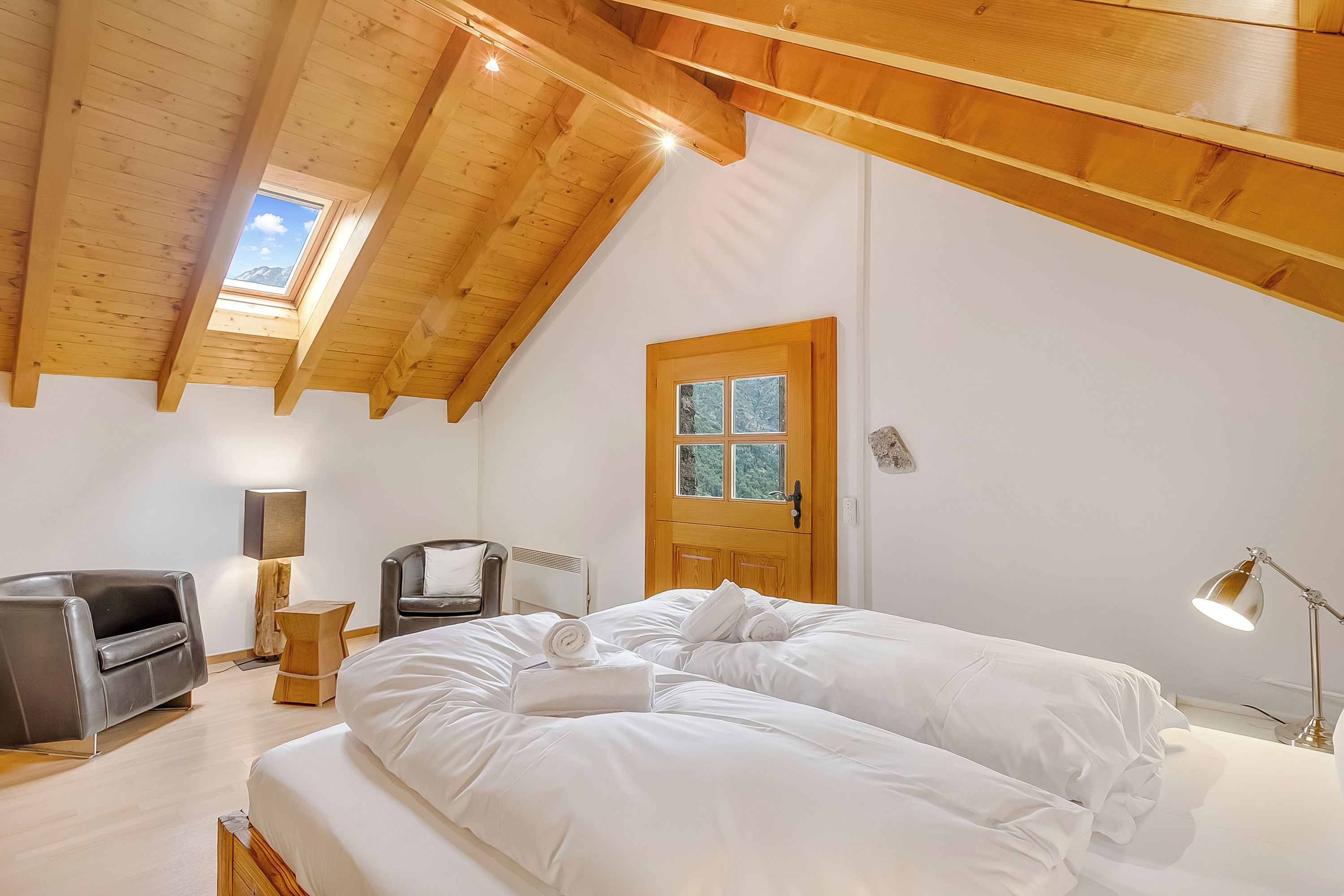 A bedroom with a large bed, a wooden ceiling, a window, a door, and a desk with a lamp.