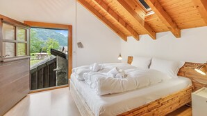 A bedroom with a wooden ceiling, a bed with white linens, a nightstand, and a window overlooking a mountainous landscape.