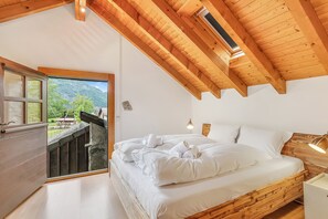 A bedroom with a wooden ceiling, a bed with white linens, a nightstand, and a window overlooking a mountainous landscape. - Casa Eyrie Centovalli 3BR Chalet (Centovalli)