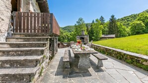 A stone staircase leading to a wooden deck with a stone table set for dining, surrounded by lush greenery and a stone oven. - Casa Eyrie Centovalli 3BR Chalet (Centovalli)