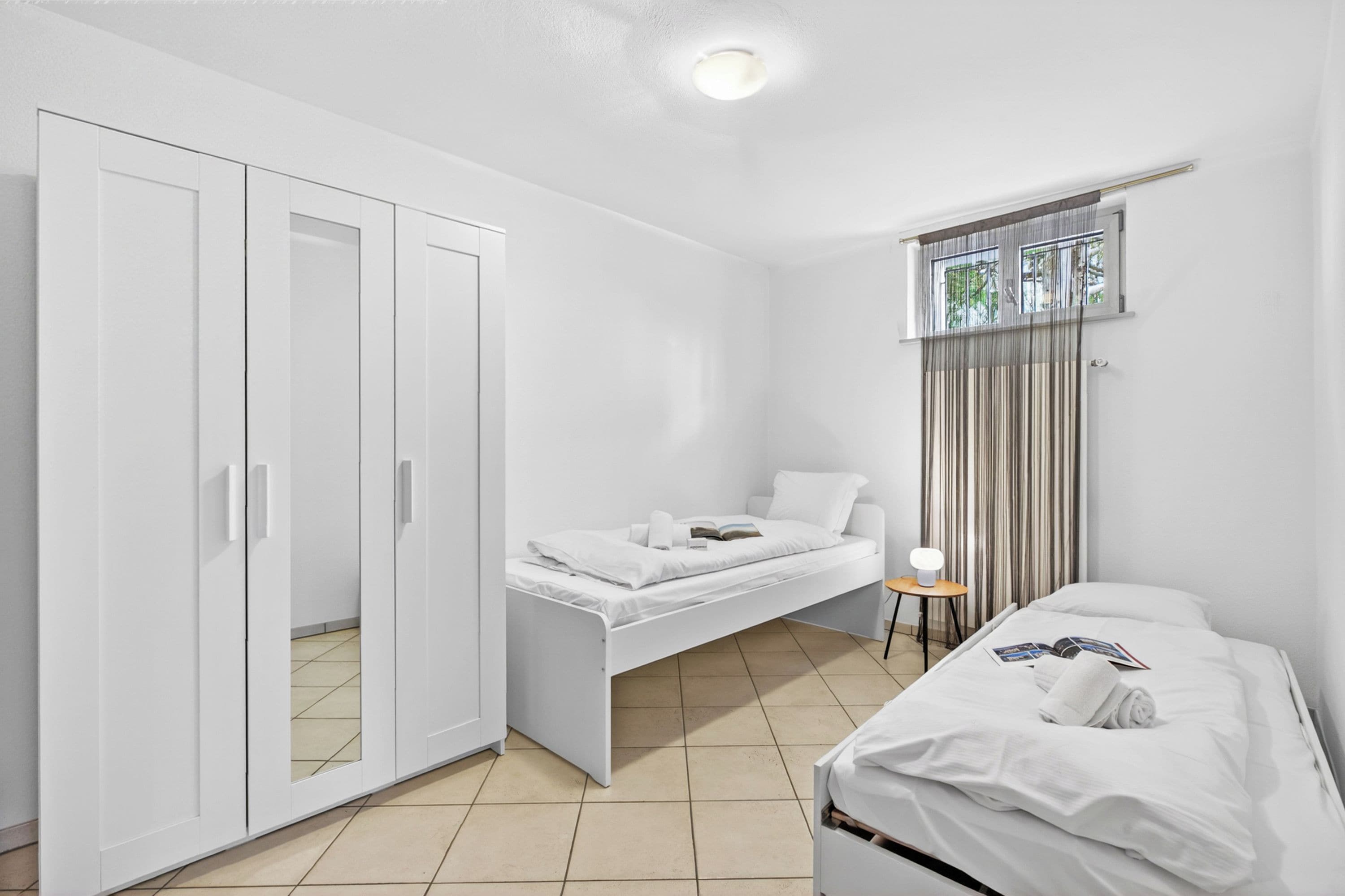 A bedroom with a bed, a wardrobe, a small table with a lamp, and a window with curtains.