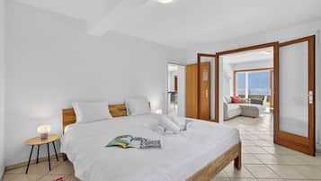 A bedroom with a large bed, a bedside table with a lamp, a view of the balcony, and a door leading to another room.