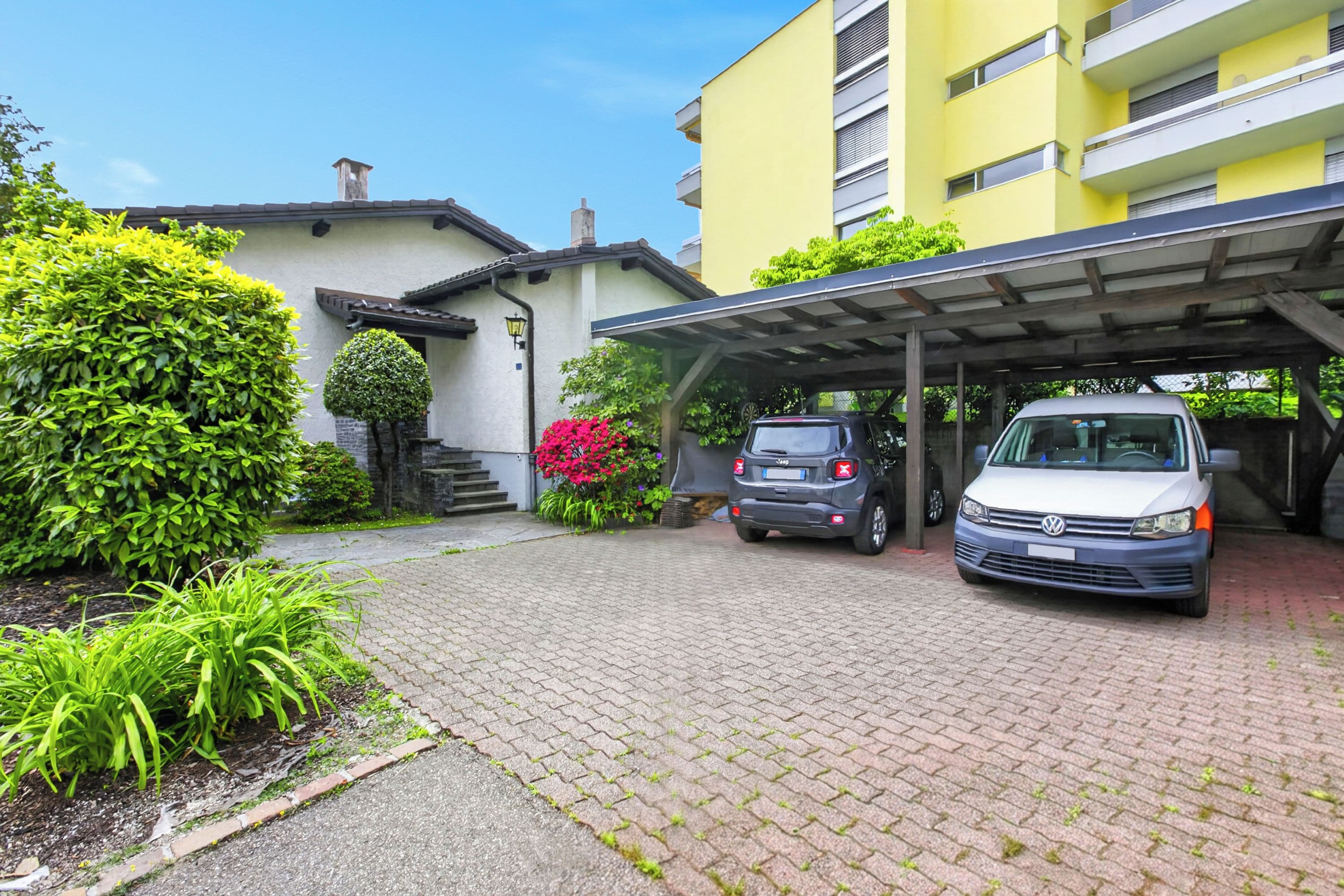 A building with a covered parking area, a white car, and a grey car parked in front.