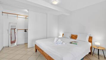 A modern bedroom with a wooden bed frame, white bedding, a bedside table with a lamp, and a door leading to another room.