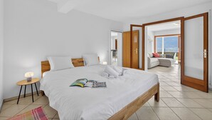 Un dormitorio con una cama grande, una mesita de noche con lámpara, vista al balcón y una puerta que da a otra habitación.