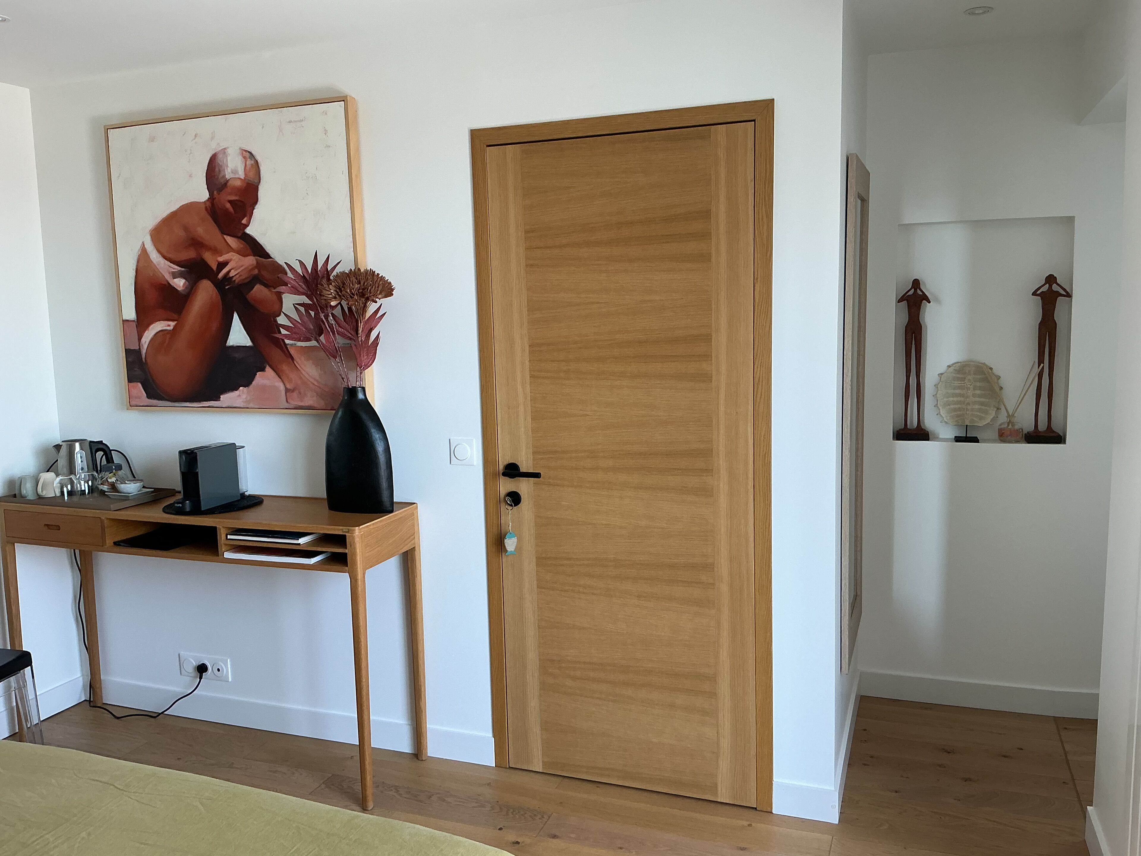 Habitació amb una porta de fusta, un escriptori amb un vas, un quadre i una estant amb objectes decoratius.
