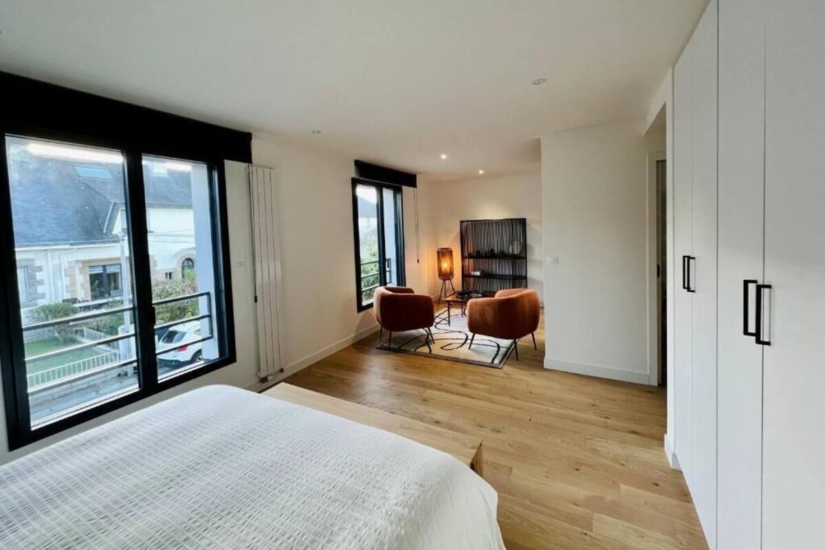 Une chambre moderne avec un grand lit, un coin salon avec deux fauteuils et une vue dégagée grâce à de grandes fenêtres.