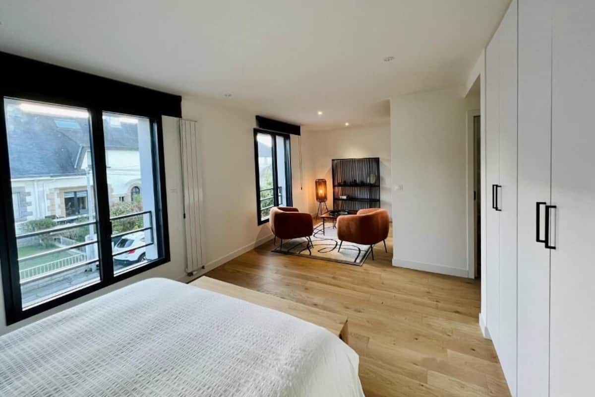 A modern bedroom with a large bed, a seating area with two chairs, and a view through large windows.