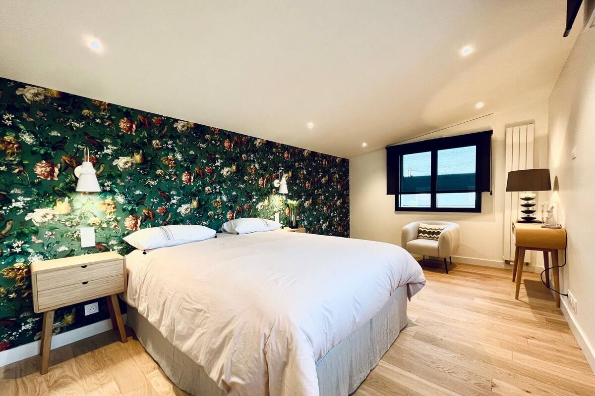 Une chambre à coucher avec un papier peint à motifs floraux, un grand lit, des tables de chevet, une chaise et une fenêtre donnant sur un beau paysage.