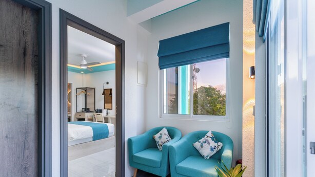 Une chambre d’hôtel moderne, équipée d’un lit, de deux fauteuils turquoise, d’une fenêtre donnant sur un paysage agréable et d’une porte menant à une autre pièce.