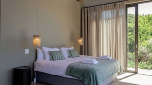 Een slaapkamer met een bed, kussens, een deken, een nachtkastje, een lamp en uitzicht op groen door een raam.