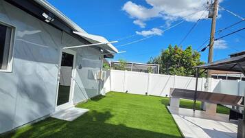 Patio trasero residencial con césped artificial, una cerca blanca y una casa con garaje.