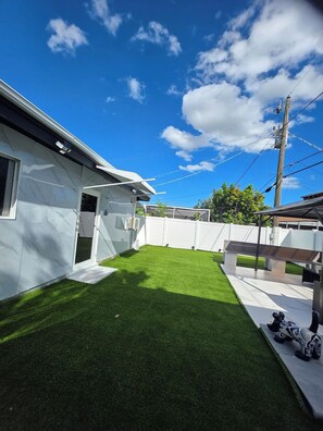 Patio trasero residencial con césped artificial, una cerca blanca y una casa con garaje.