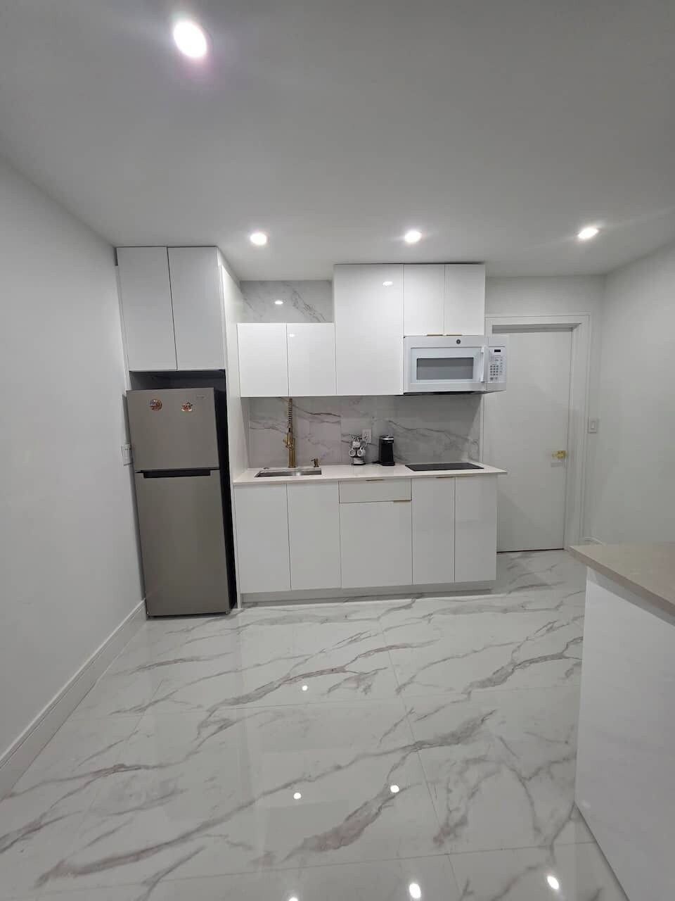 A compact kitchen with white cabinets, a stainless steel refrigerator, a microwave, and a marble floor.