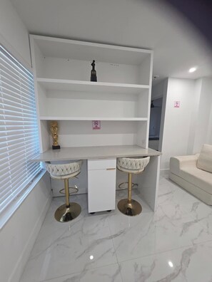 Una habitación moderna y minimalista con un escritorio blanco y dos taburetes altos, un sofá blanco y una ventana con persianas.