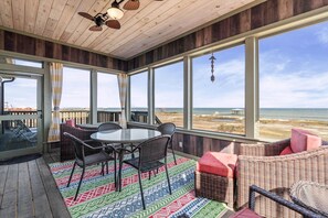 Ein überdachter Außenbereich mit Tisch und Stühlen, großen Fenstern und Blick auf den Strand.