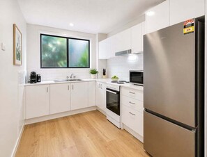 A modern kitchen with white cabinets, a stainless steel refrigerator, a microwave, and a window with a view of greenery. - Relaxed Coastal Stay in Burleigh Heads (Gold Coast)