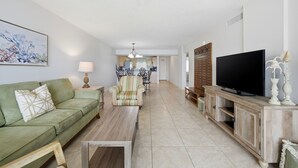 A living room with a green sofa, a wooden coffee table, a striped armchair, a TV on a wooden stand, and a kitchen in the background. - Welcome to Sandy Key 537 (Perdido Key)