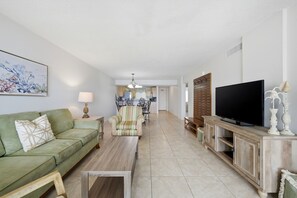 A living room with a green sofa, a wooden coffee table, a striped armchair, a TV on a wooden stand, and a kitchen in the background. - Welcome to Sandy Key 537 (Perdido Key)