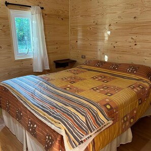 Ein Zimmer einer Holzhütte mit einem Bett, einem Fenster mit Vorhängen und einem kleinen Nachttisch aus Holz.