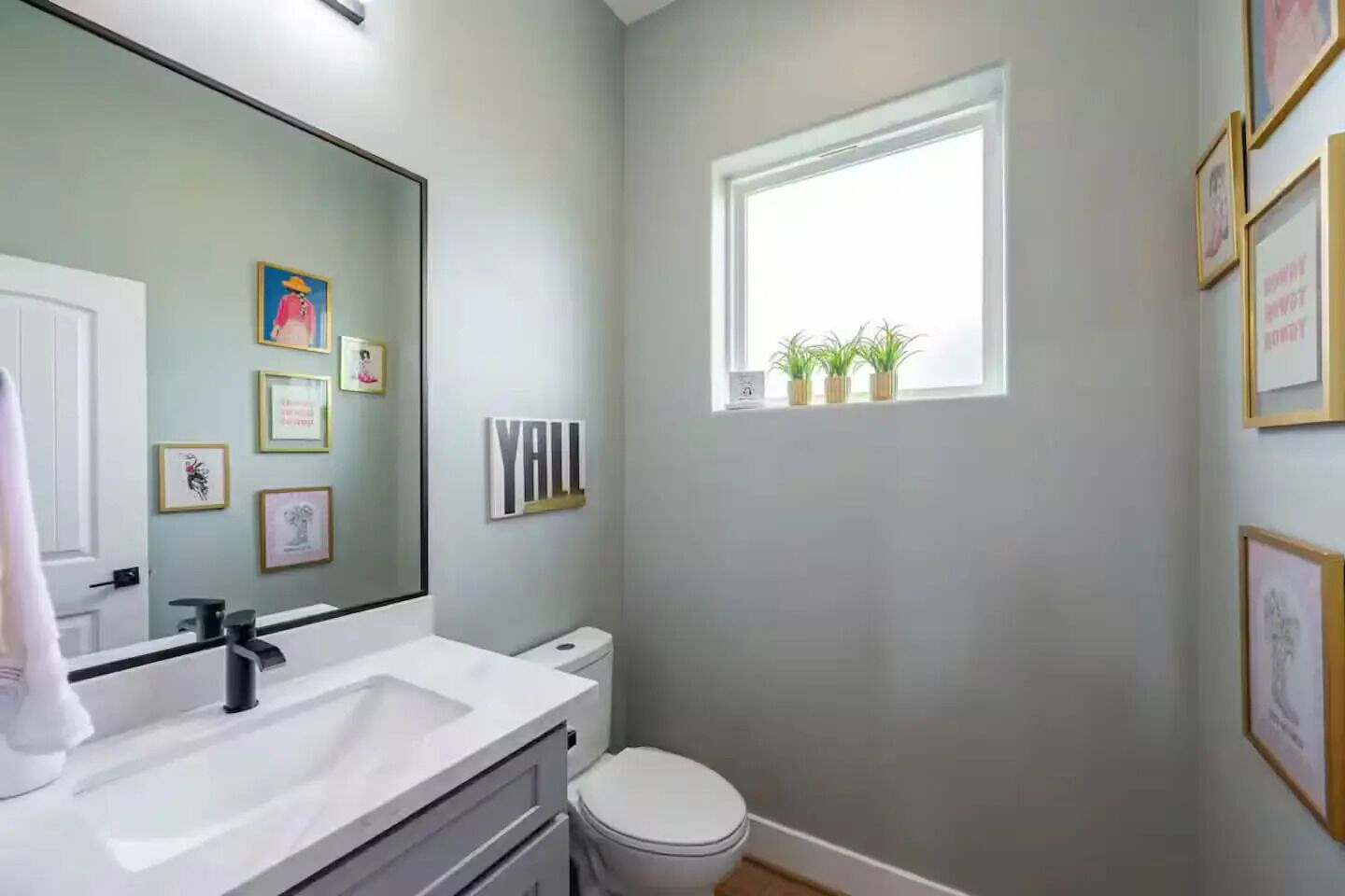 Un baño con un lavabo blanco, un inodoro, un espejo grande, obras de arte enmarcadas en las paredes y una ventana con plantas.