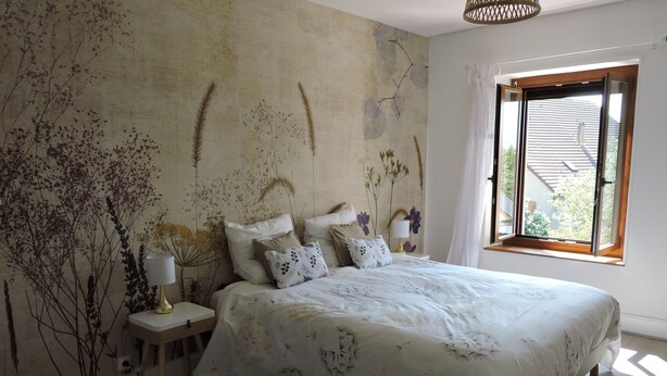 A bedroom with a large bed, a nightstand with a lamp, a wall mural, and a window with curtains.