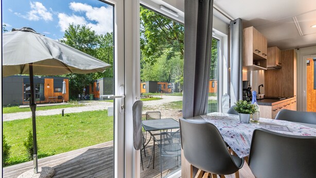 A modern kitchen with wooden cabinets, a dining area with a table and chairs, and a view of a grassy area and other buildings.
