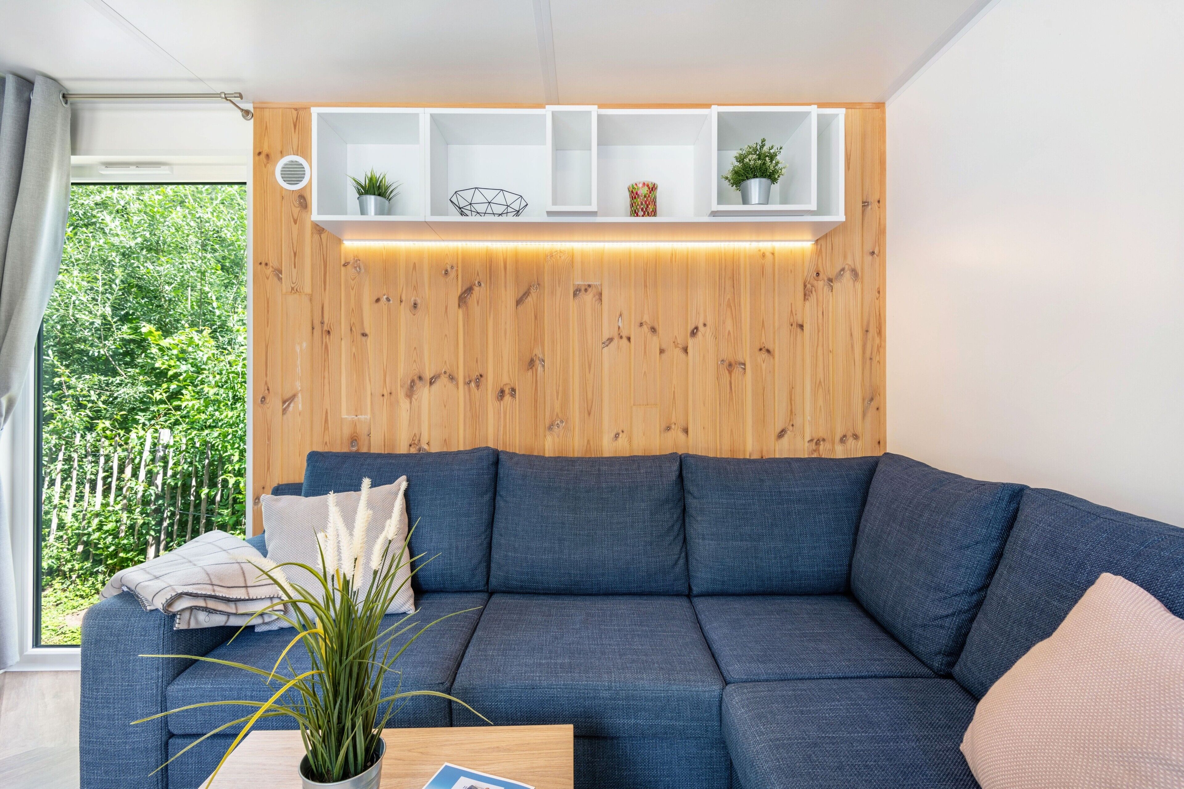 En moderne stue med en blå sofa, træpaneler og et vindue med udsigt til grøn vegetation.