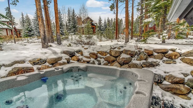 Un bain à remous entouré de terre et d’arbres recouverts de neige, avec un mur de pierre et une maison en arrière-plan.