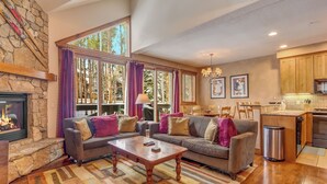 Un salon doté d’une cheminée en pierre, de grandes fenêtres ornées de rideaux violets, d’un ensemble de canapés et d’un coin cuisine.