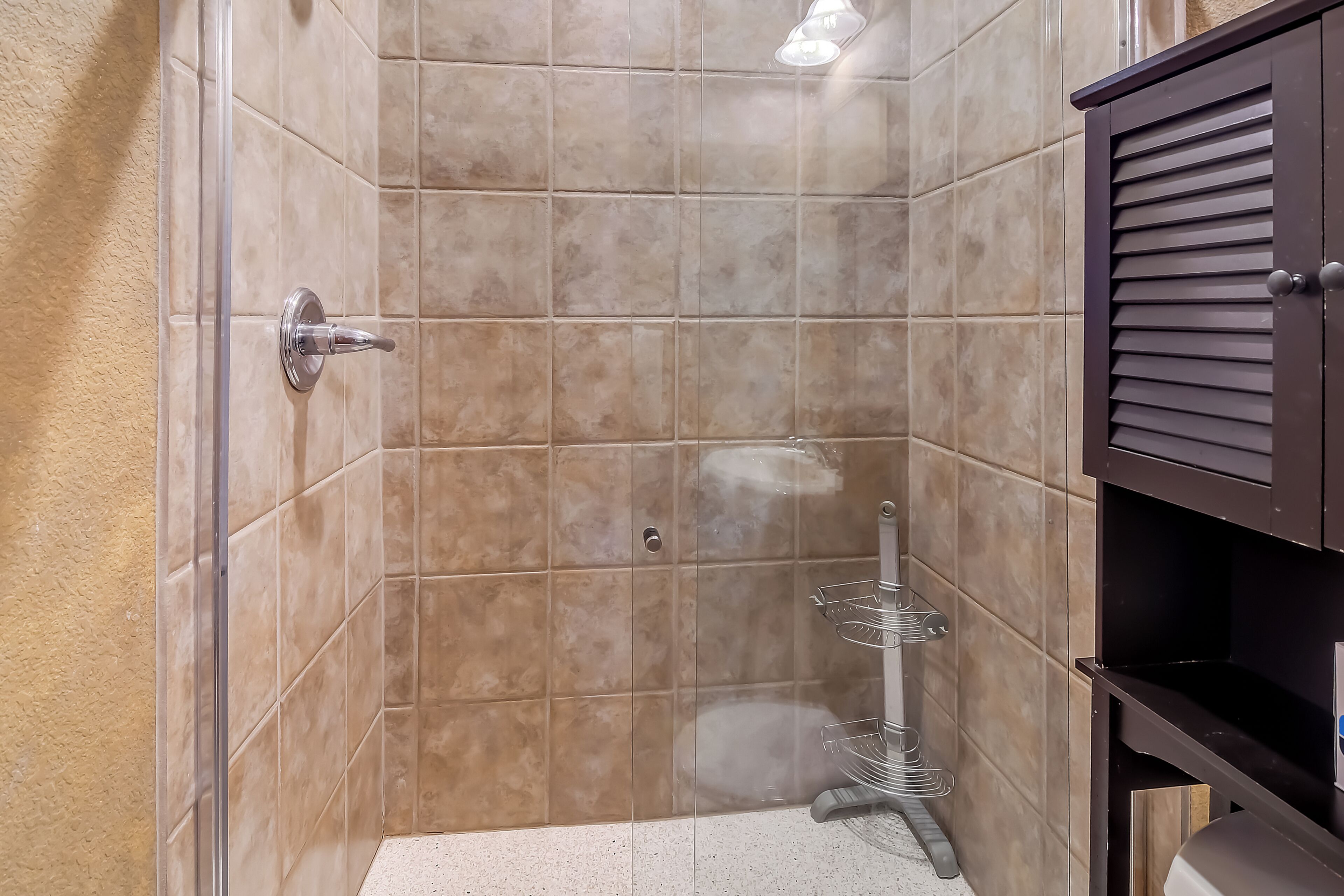 Une douche avec une porte en verre, des carreaux de couleur beige et un meuble en bois foncé.