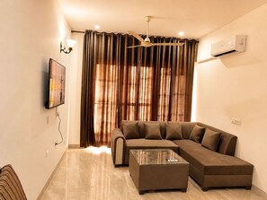 Una sala de estar con un sofá modular, una mesa de centro de vidrio, un ventilador de techo y un aire acondicionado instalado en la pared.