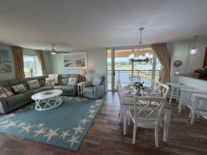 Ein Wohnzimmer mit einer Eckcouch, einem Esstisch mit Stühlen, einem kleinen Beistelltisch, einem Teppich mit Sternfischmuster und einem Balkon mit Blick.
