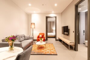 Habitación de hotel moderna con sofá, una mesita de centro, televisor, un banco y un baño visible a través de una puerta abierta.