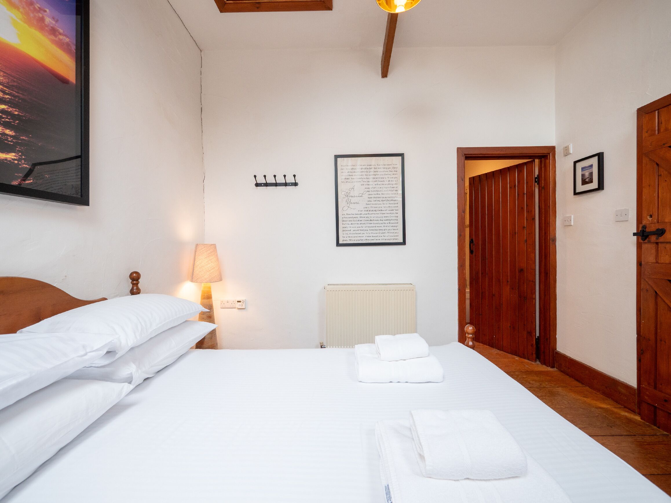 A hotel room with a bed, a lamp, a framed poem, a coat rack, and a wooden door.