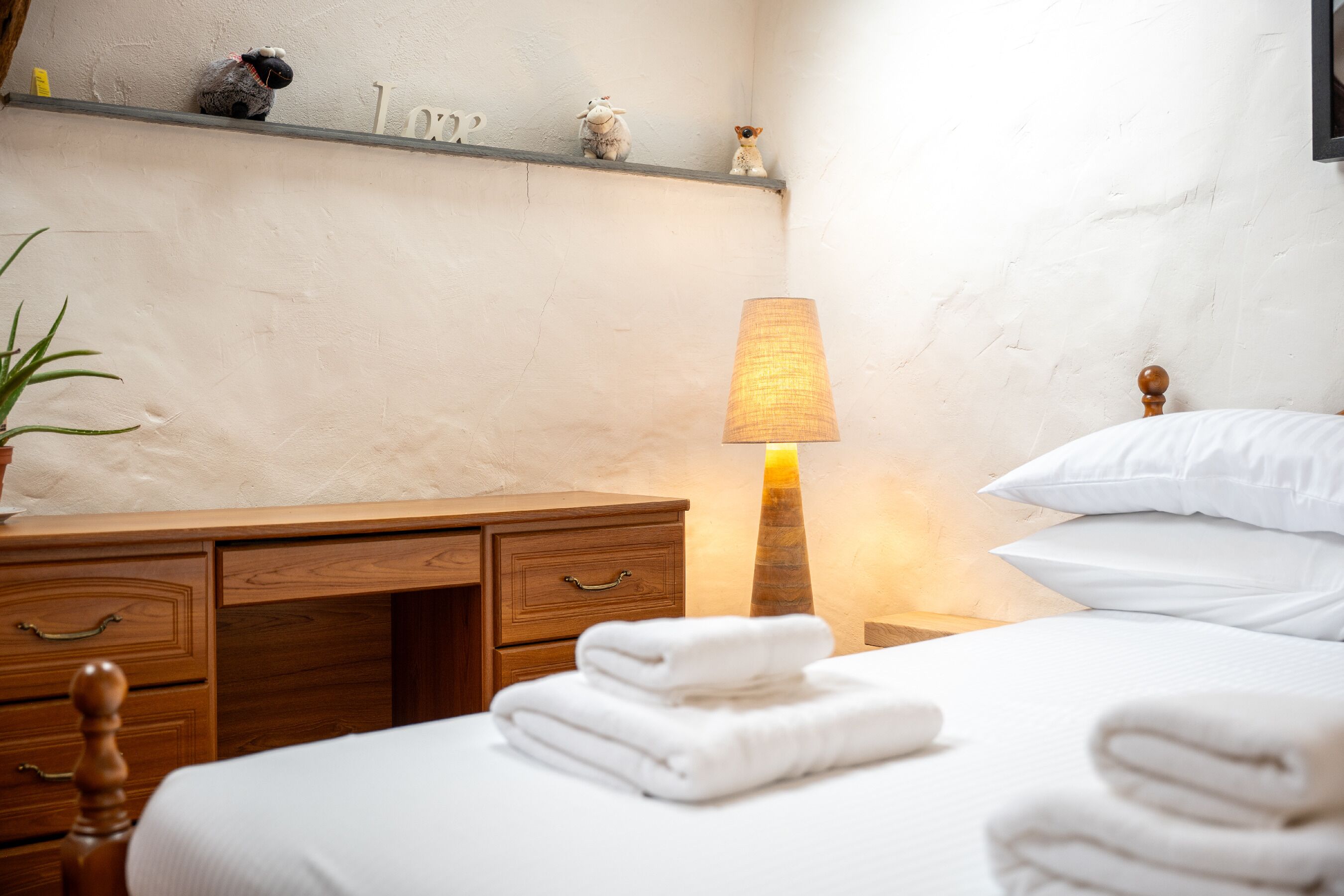 A neatly made bed with folded white towels, a wooden nightstand with a lamp, a wooden dresser with drawers, and a shelf with decorative items.